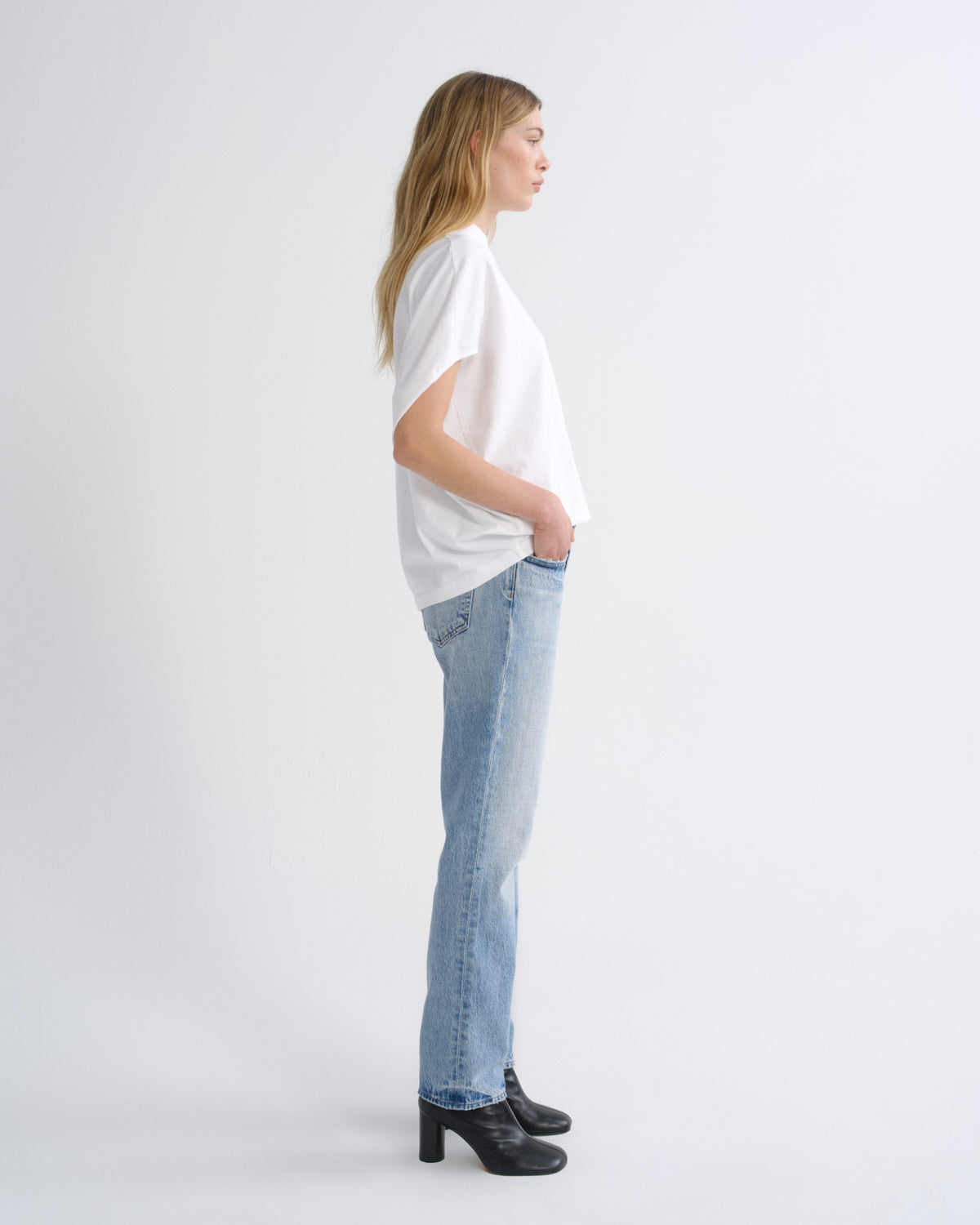 A woman in profile wearing a white short-sleeve t-shirt, light-wash blue jeans, and black high-heeled boots, standing against a plain light gray studio background.