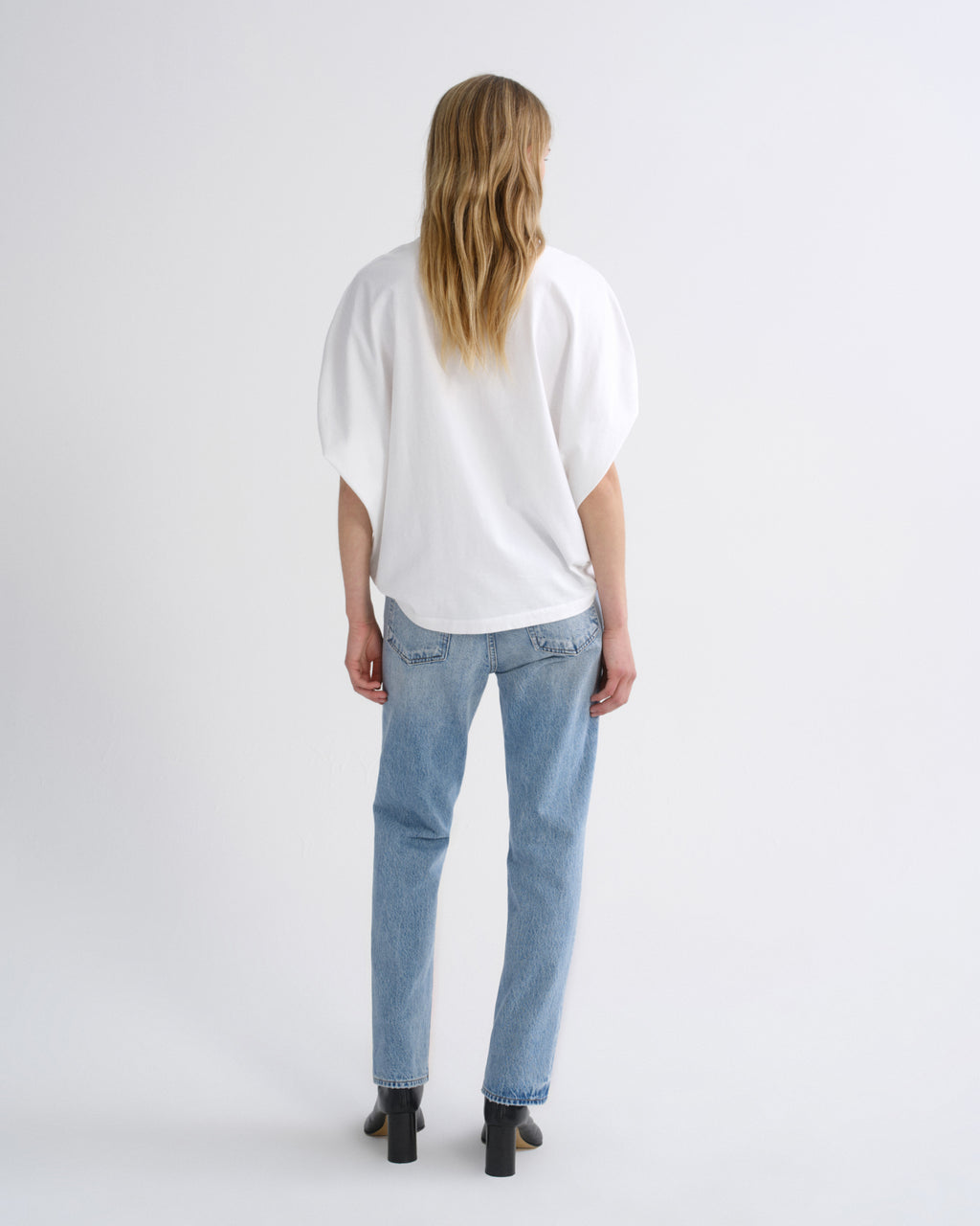 A woman viewed from behind wearing a loose white short-sleeve top, light blue jeans, and black block-heel boots against a plain light gray studio background.
