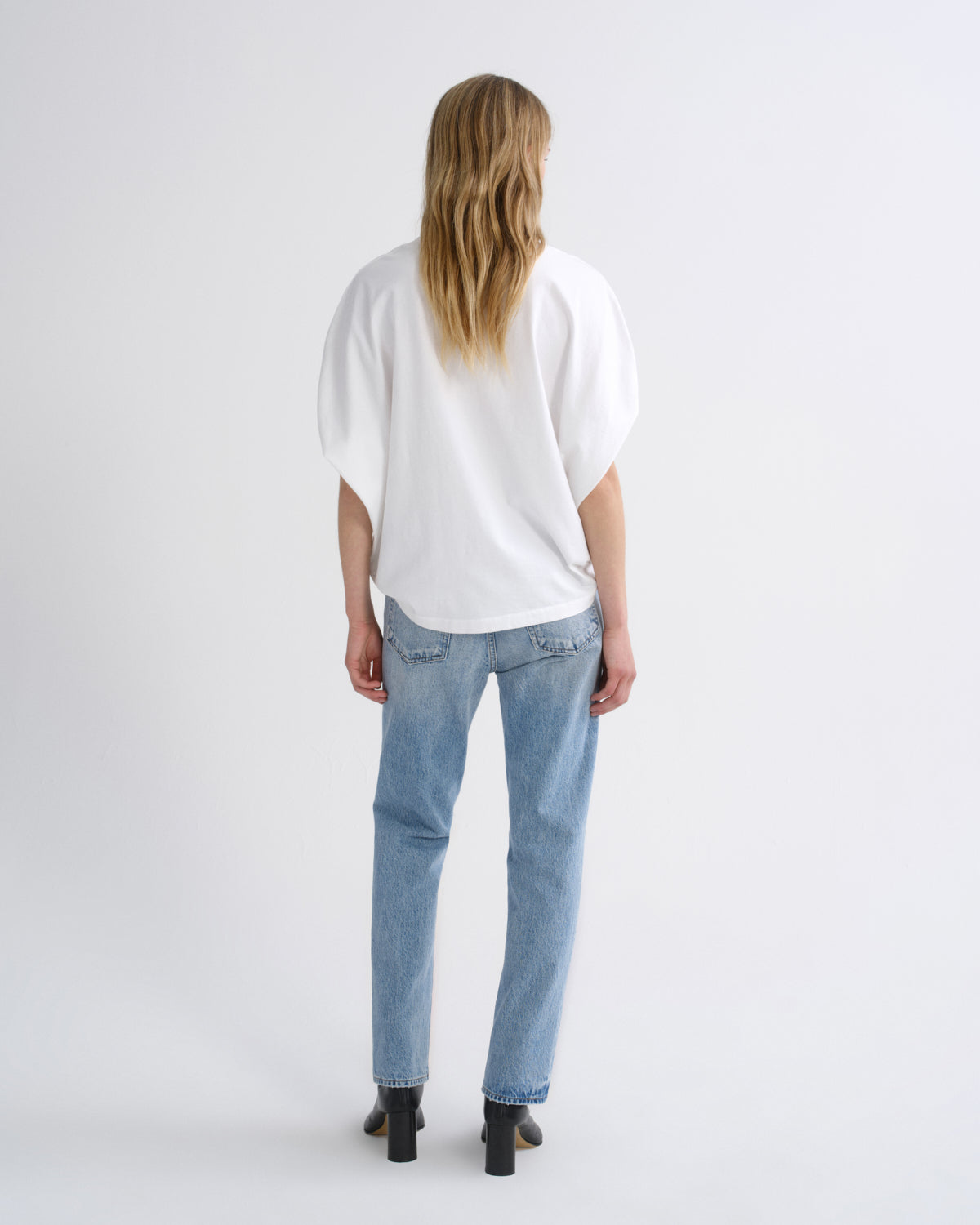 A woman viewed from behind wearing a loose white short-sleeve top, light blue jeans, and black block-heel boots against a plain light gray studio background.