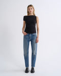 A woman modeling a black off-the-shoulder short-sleeve top and blue jeans against a plain light gray studio background.