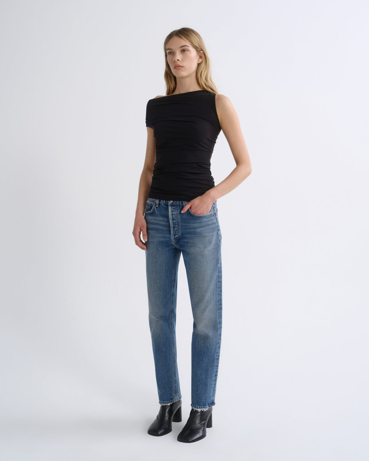 A woman modeling a black sleeveless top and blue jeans with black ankle boots, standing against a plain light gray studio background.