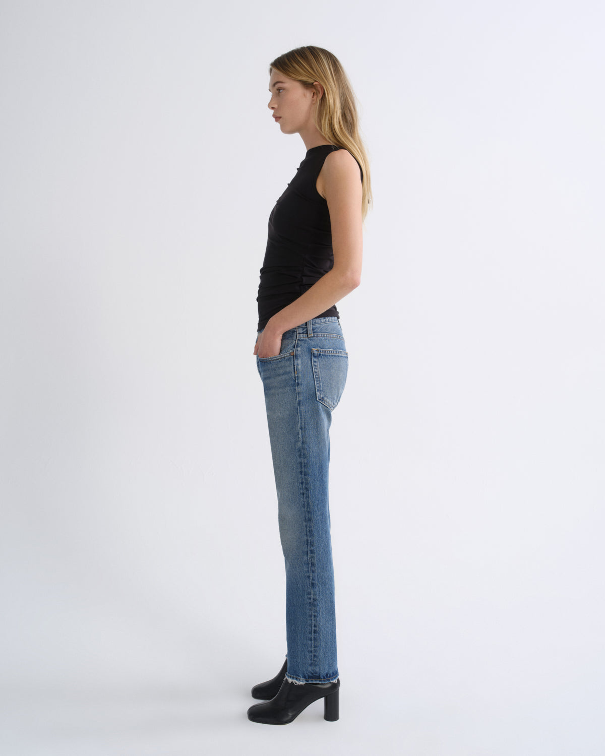 A woman in a black sleeveless top and light blue jeans stands in profile against a plain light gray studio background, wearing black heeled boots.