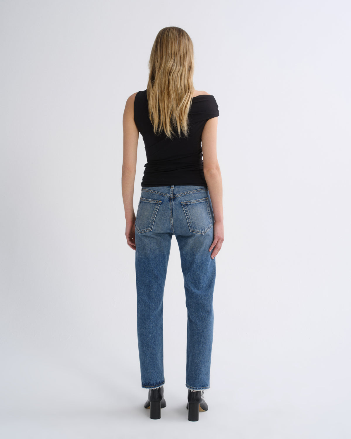 A woman viewed from behind wearing a black off-the-shoulder top, blue jeans, and black ankle boots against a plain light gray studio background.