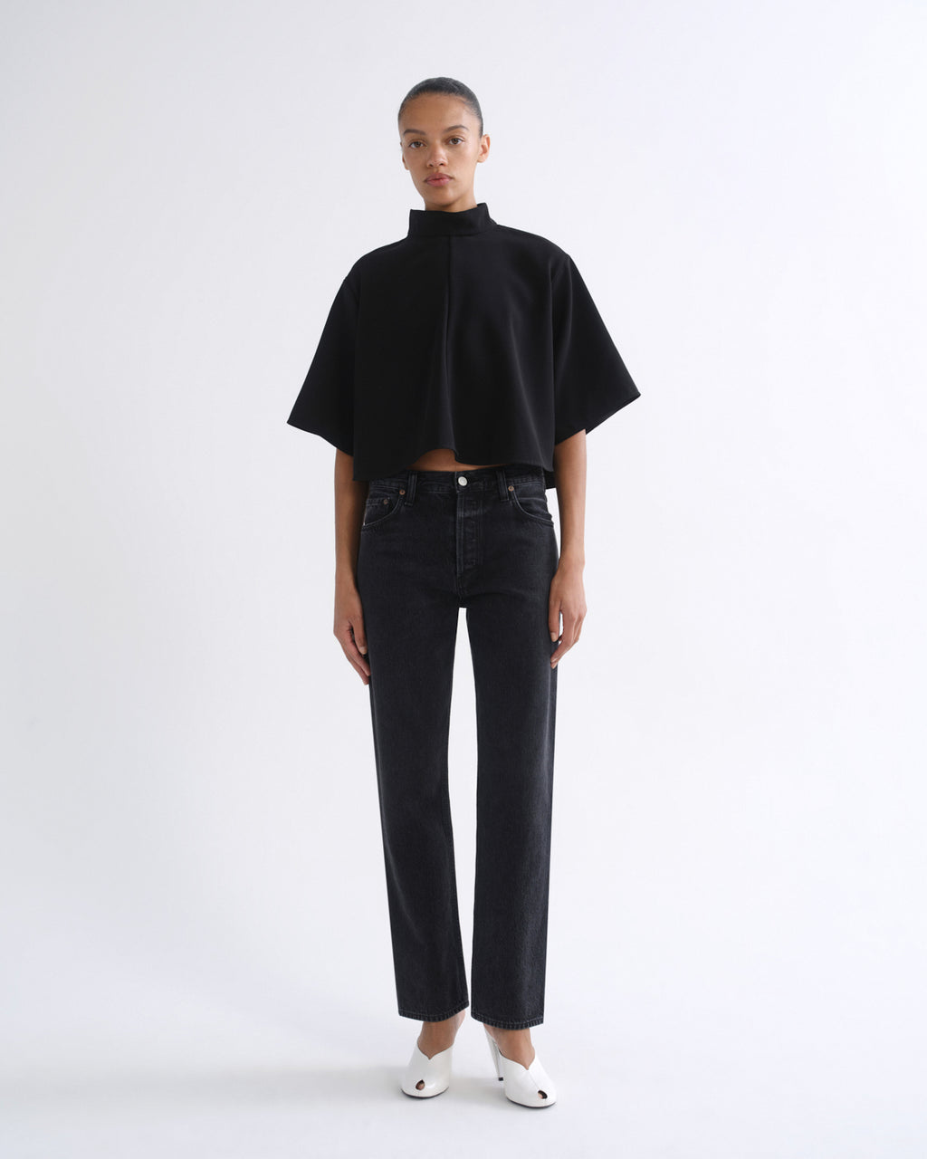 A model stands against a plain white studio background wearing a black cropped short-sleeve top with a high mock neck, black jeans, and white pointed shoes.