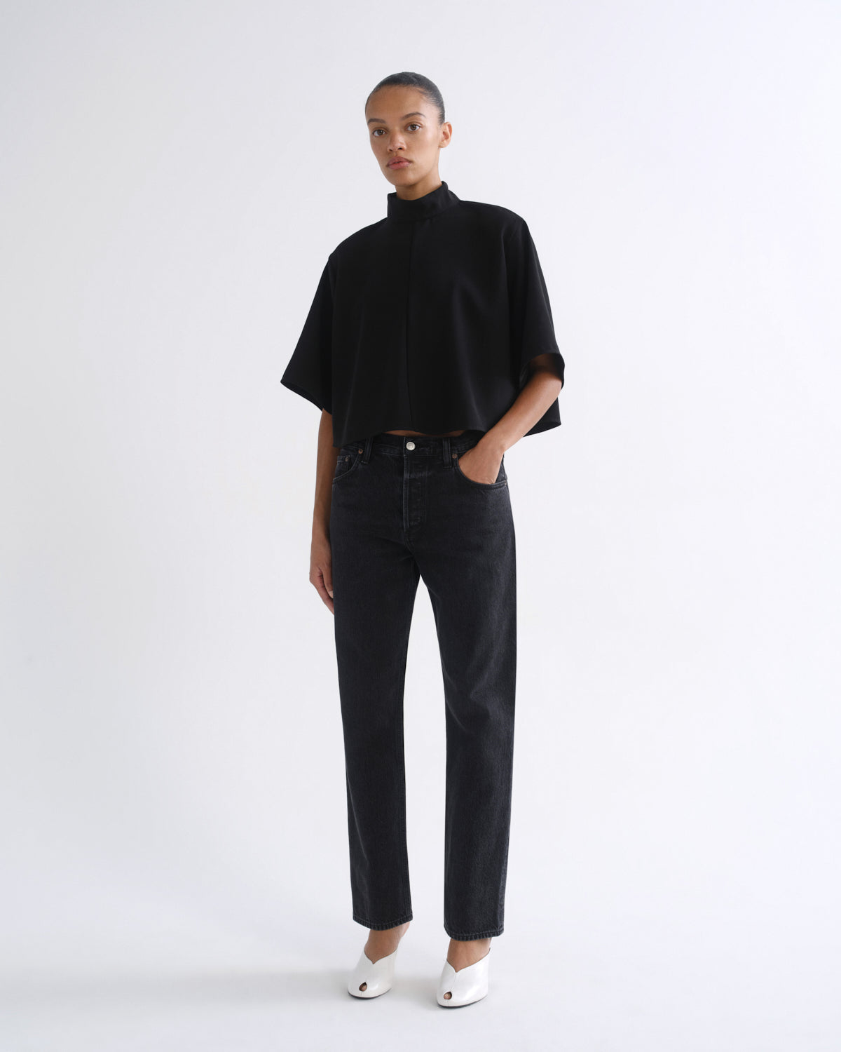 A model stands against a plain white studio background wearing a black short-sleeve mock-neck top, black straight-leg jeans, and white pointed shoes.