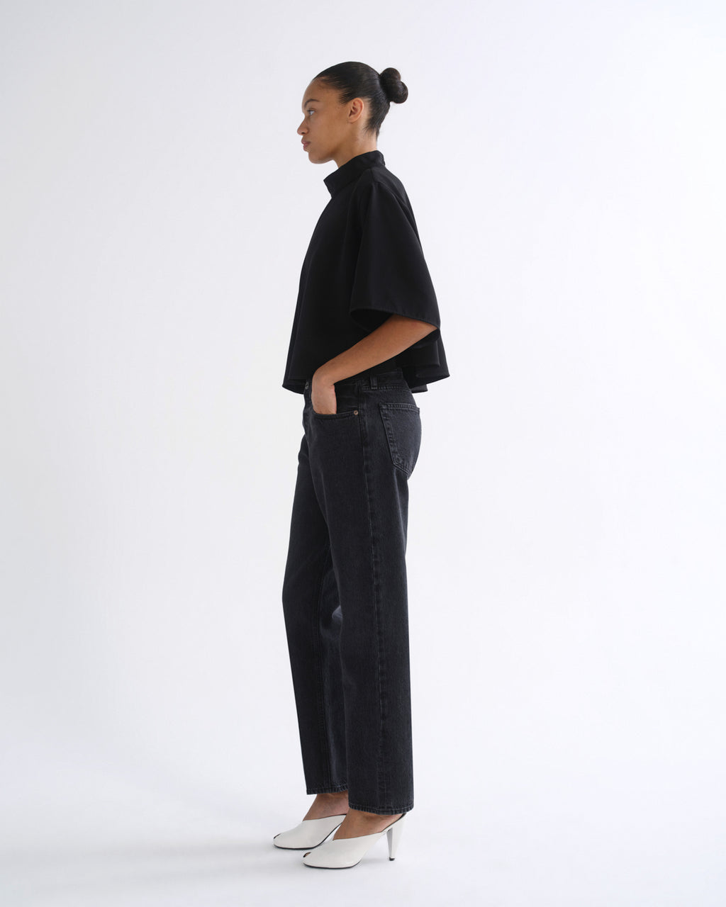 A model shown in profile wearing a black short-sleeve mock-neck top, black wide-leg jeans, and white pointed-toe heels against a plain white studio background.
