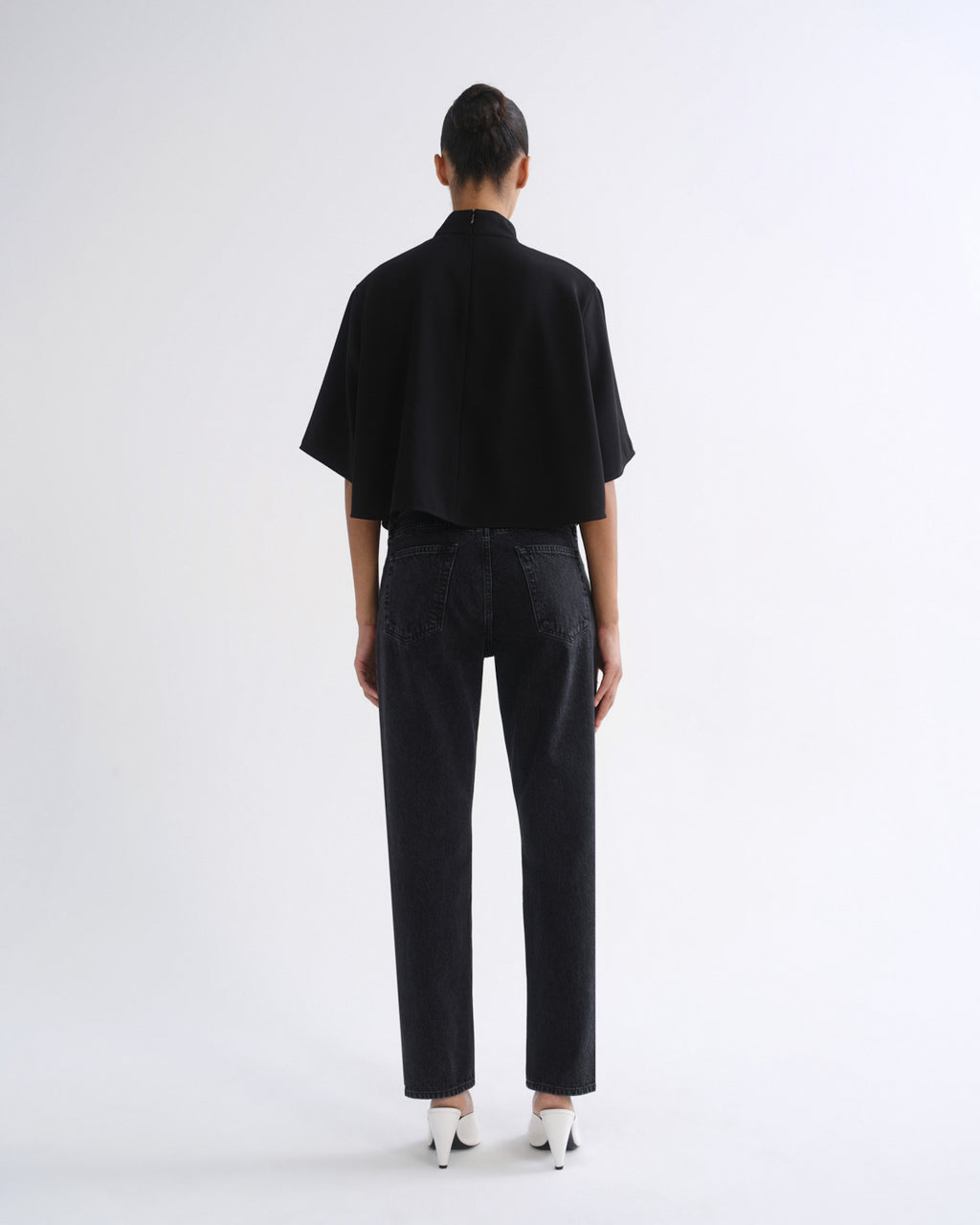 A model viewed from the back wearing a black short-sleeved high-neck top, black wide-leg jeans, and white pointed-toe heels against a plain white studio background.