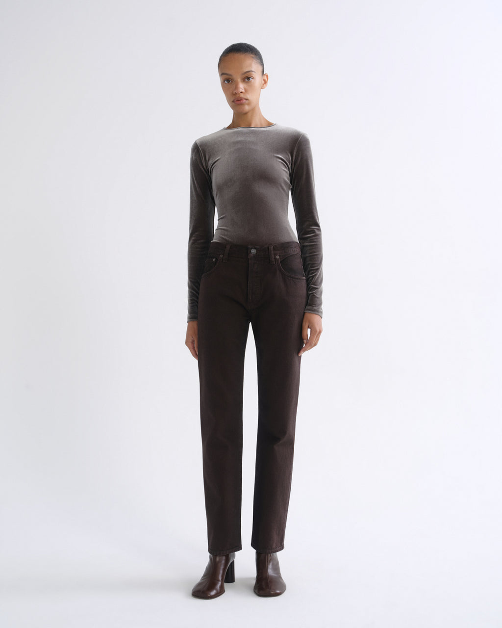 A model stands against a light gray studio background wearing a fitted long-sleeve taupe top, dark brown straight-leg jeans, and dark brown ankle boots.