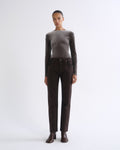 A model stands against a light gray studio background wearing a fitted long-sleeve taupe top, dark brown straight-leg jeans, and dark brown ankle boots.
