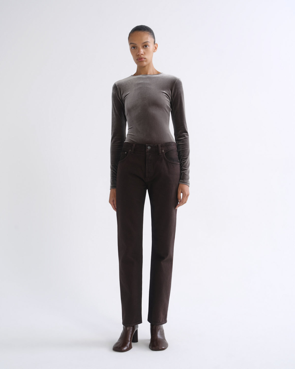 A model stands against a light gray studio background wearing a fitted long-sleeve taupe top, dark brown straight-leg jeans, and dark brown ankle boots.