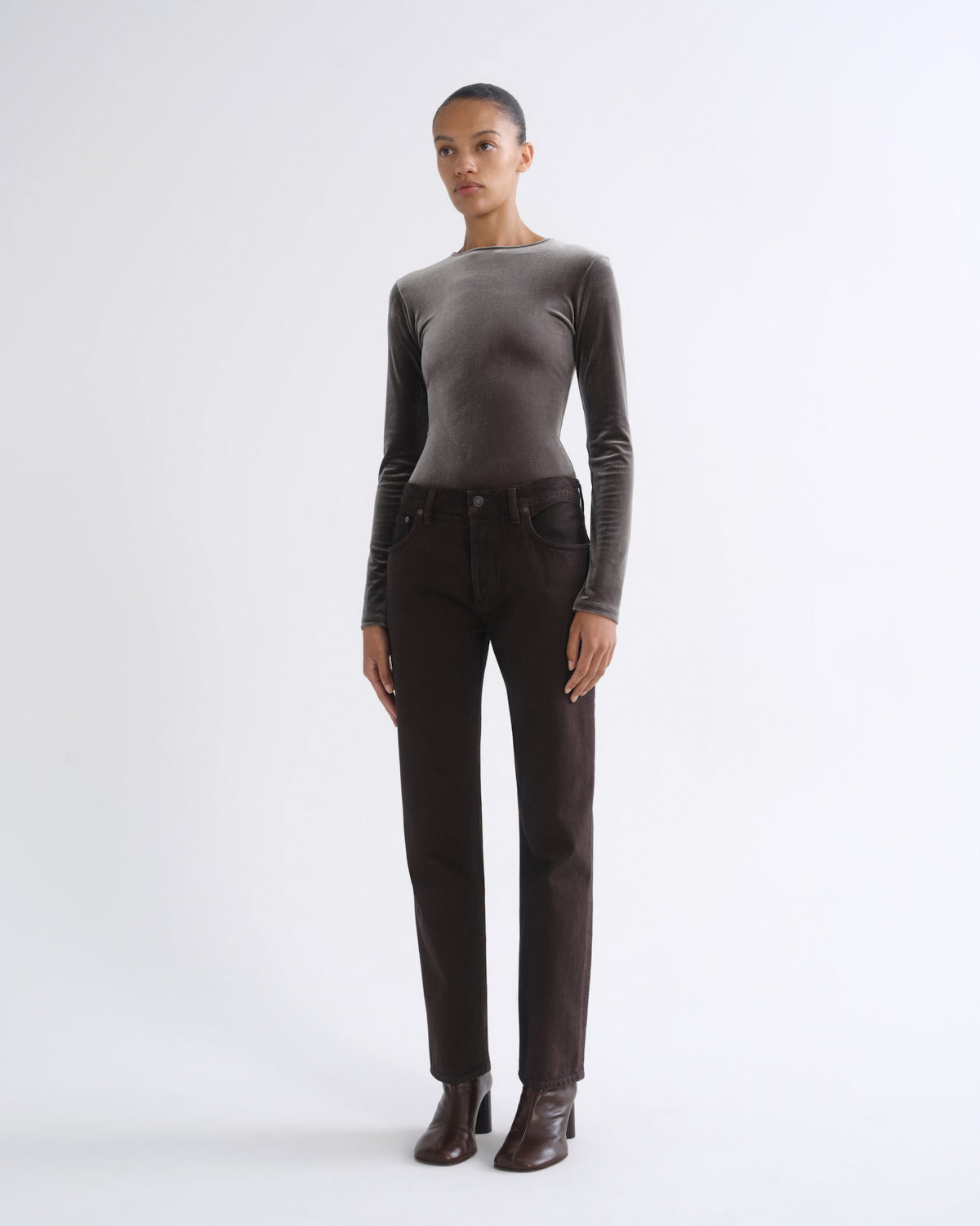 A model stands against a light gray studio background wearing a dark brown long-sleeve velvet top, dark brown straight-leg jeans, and dark brown heeled ankle boots.