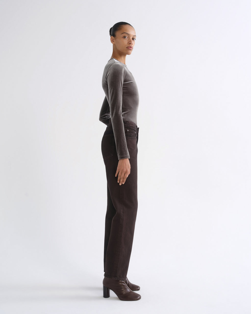A model stands in profile wearing a fitted long-sleeve velvet top in taupe-brown, paired with dark brown trousers and matching brown heeled ankle boots, against a clean white studio background.