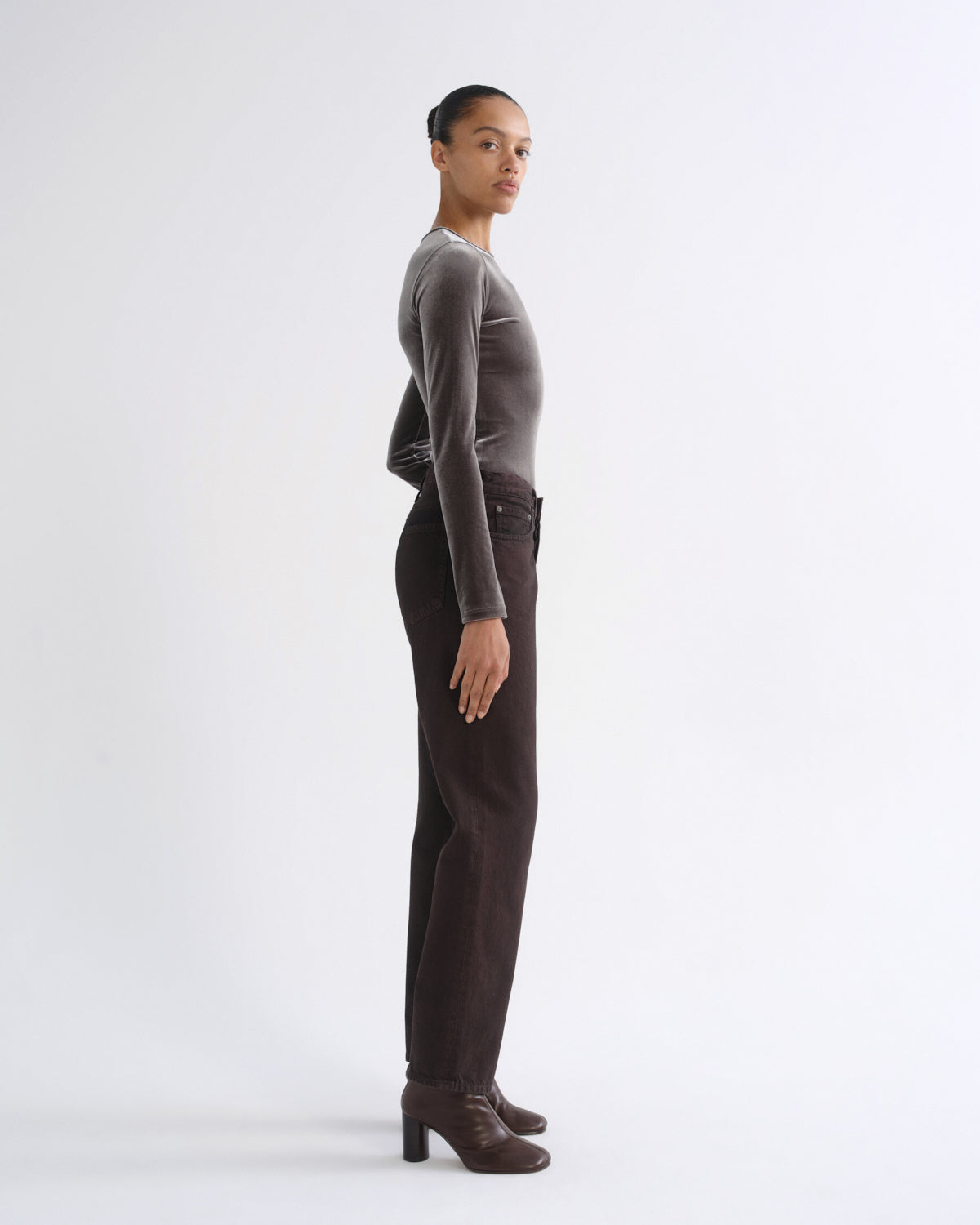 A model stands in profile wearing a fitted long-sleeve velvet top in taupe-brown, paired with dark brown trousers and matching brown heeled ankle boots, against a clean white studio background.