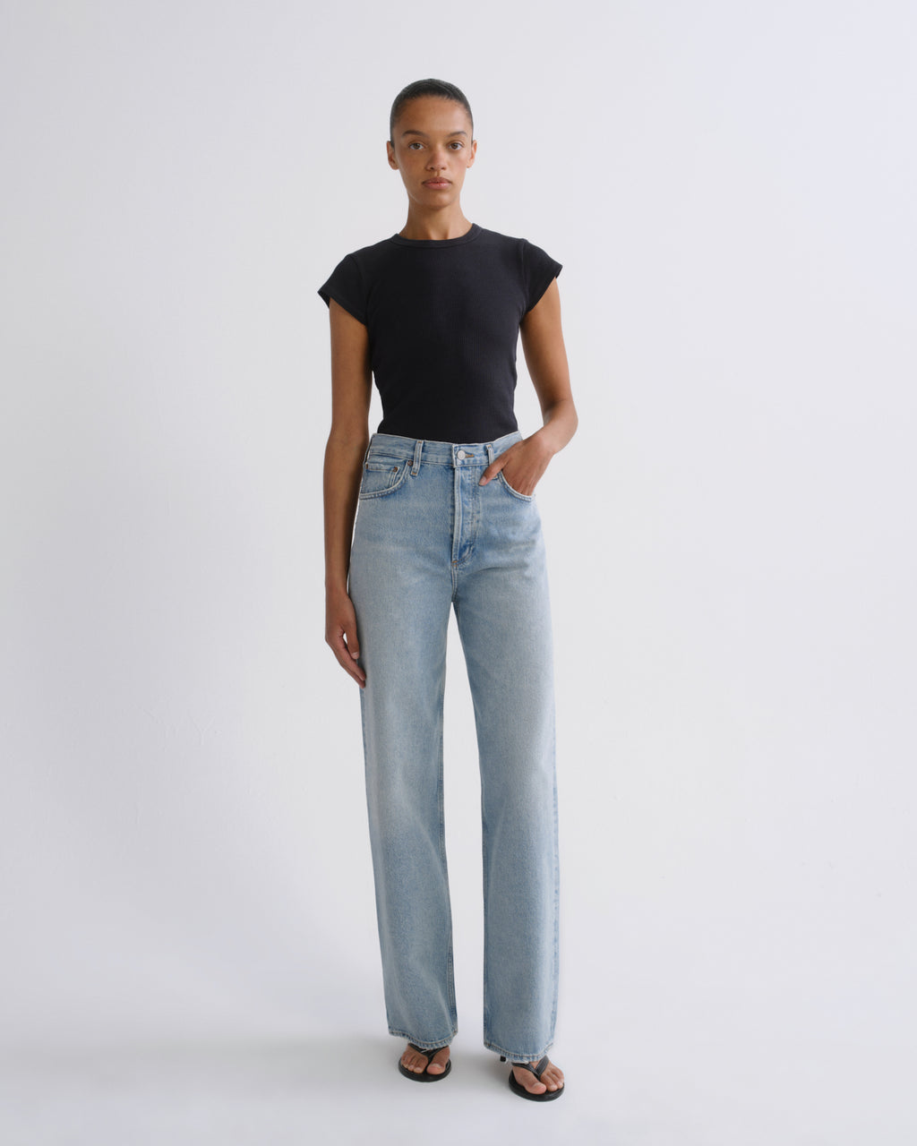 A model stands against a light gray studio background wearing a black short-sleeve crewneck top and light-wash wide-leg jeans.