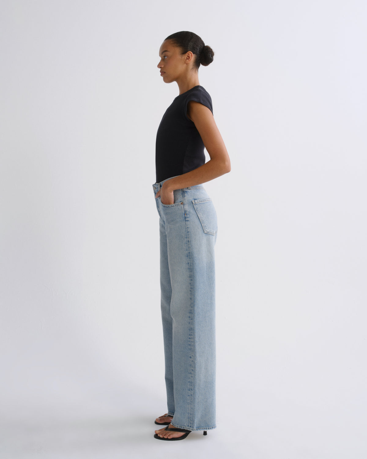 A woman in a black sleeveless top and light-wash wide-leg jeans stands in profile against a plain light gray studio background.