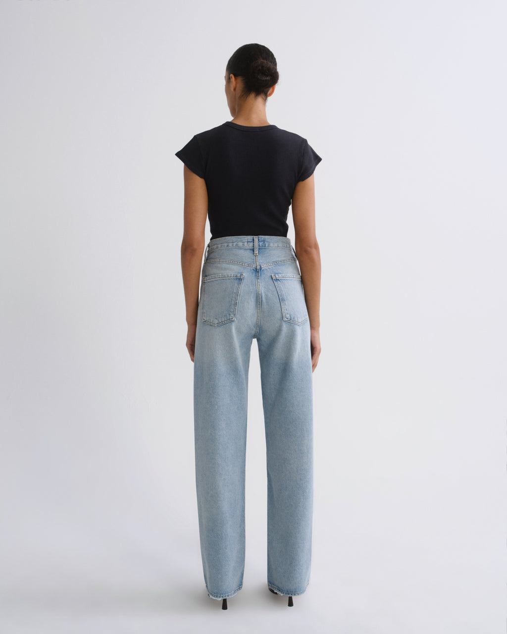 A model viewed from behind wearing a black short-sleeve crop top and light-wash wide-leg jeans against a plain light gray studio background.