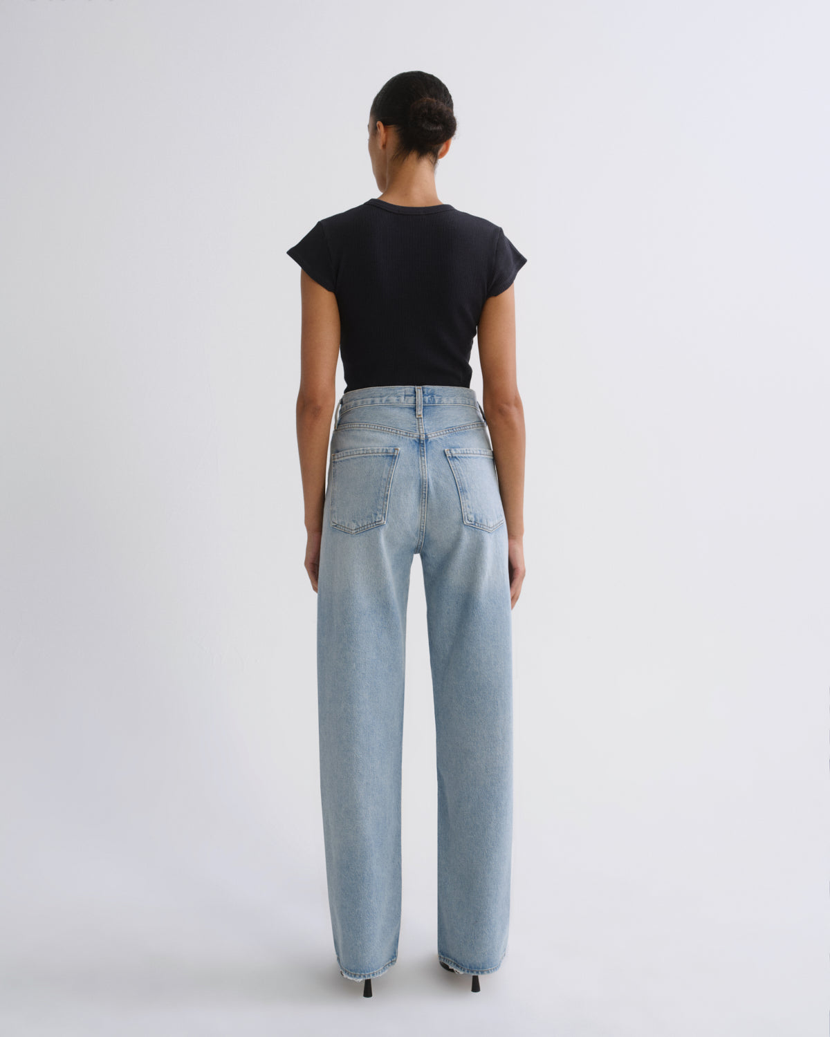 A model viewed from behind wearing a black short-sleeve crop top and light-wash wide-leg jeans against a plain light gray studio background.