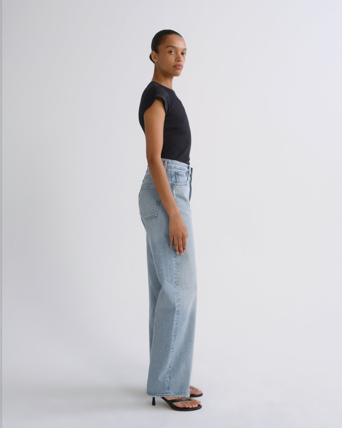 A model stands in profile wearing a black short-sleeve top, light-wash wide-leg denim jeans, and black heeled sandals against a plain light gray studio background.