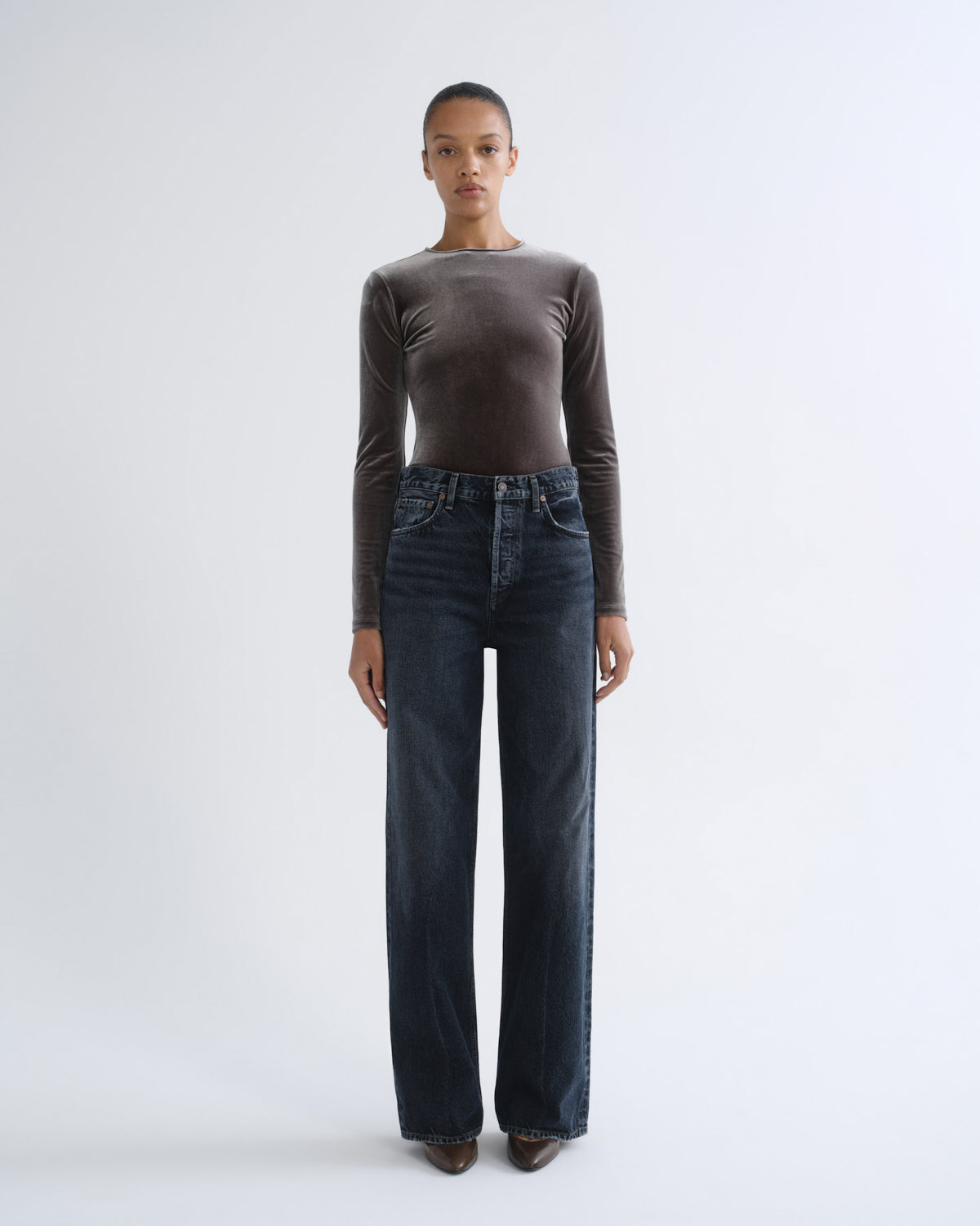 A model stands against a light gray studio background wearing a dark brown long-sleeve velvet top and dark blue wide-leg jeans.