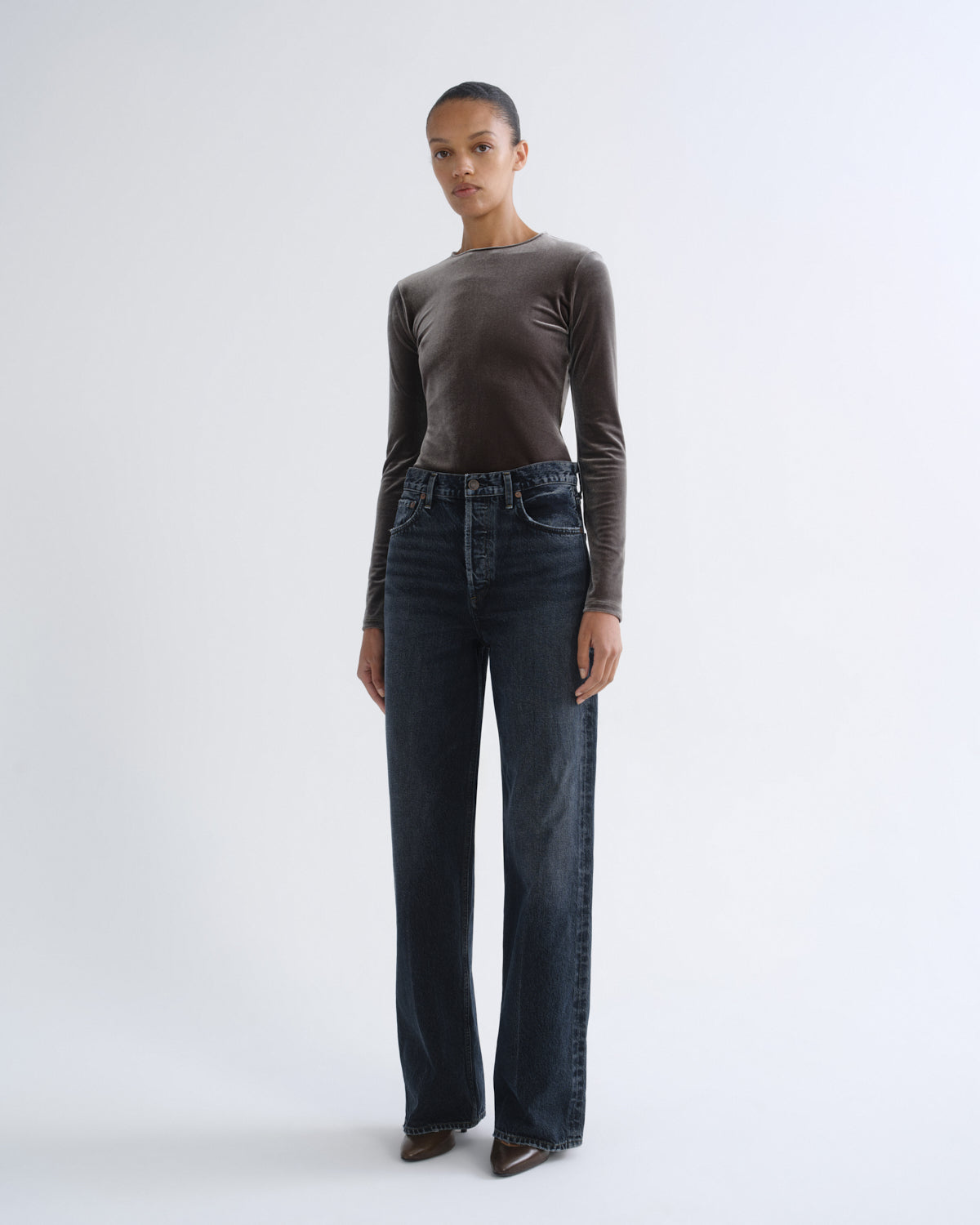 A model stands against a light gray studio background wearing a dark brown long-sleeve knit top and dark blue wide-leg jeans.