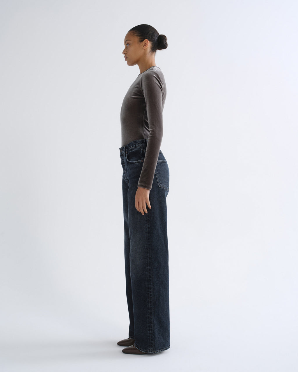 A model standing in profile wearing a dark brown long-sleeve top and dark blue wide-leg jeans against a plain white studio background.