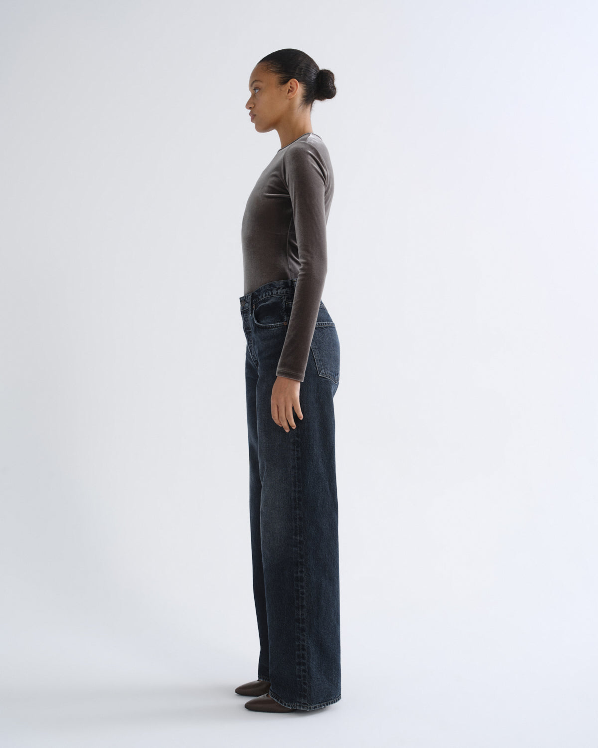 A model standing in profile wearing a dark brown long-sleeve top and dark blue wide-leg jeans against a plain white studio background.