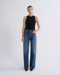 A model stands against a light gray studio background wearing a black sleeveless mock-neck top and blue wide-leg jeans.