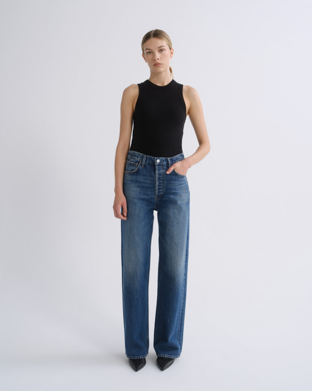 A model stands against a light gray studio background wearing a black sleeveless mock-neck top and blue wide-leg jeans.