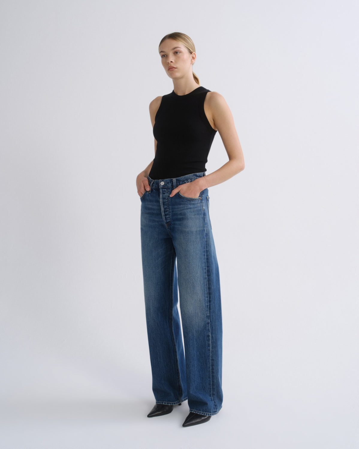 A model stands against a light gray studio background wearing a black sleeveless top, wide-leg blue jeans, and black pointed shoes.