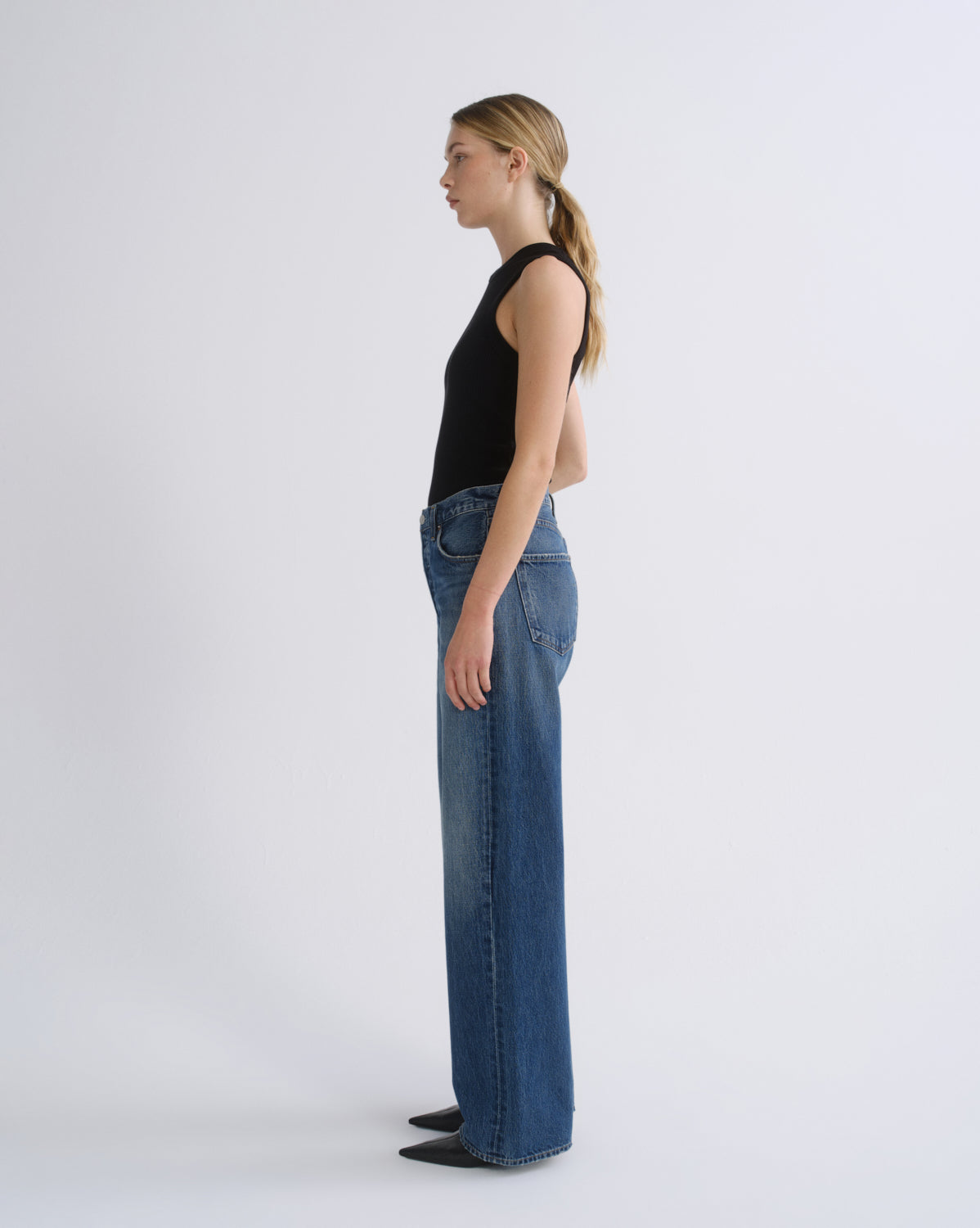A woman in profile wearing a black sleeveless top and wide-leg blue jeans, standing against a plain light gray studio background.