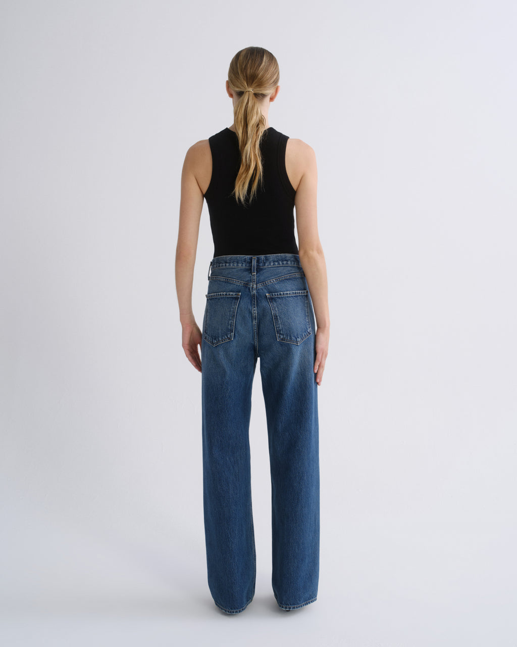 A woman viewed from the back wearing a black sleeveless top and wide-leg blue jeans against a plain light gray studio background.