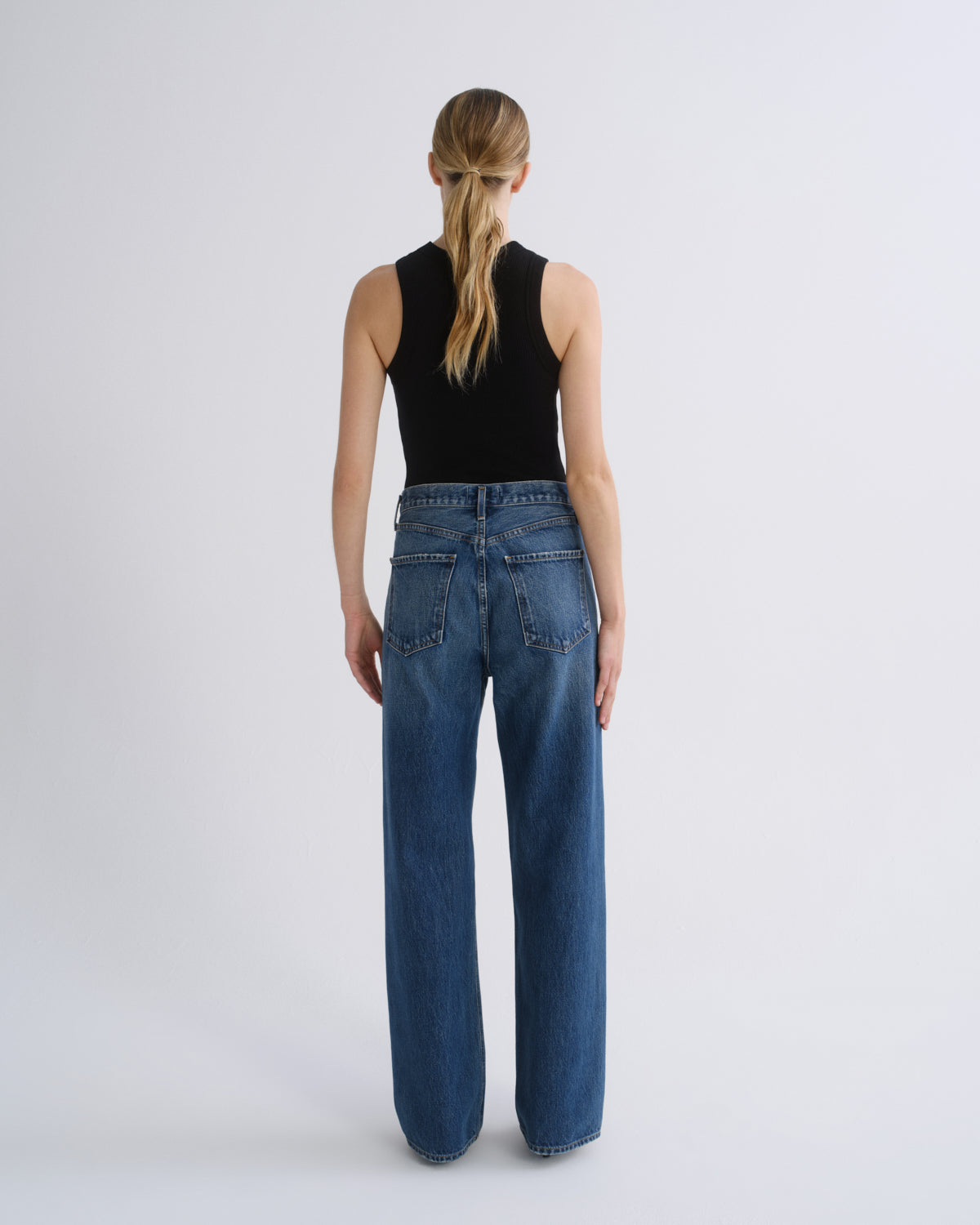 A woman viewed from the back wearing a black sleeveless top and wide-leg blue jeans against a plain light gray studio background.