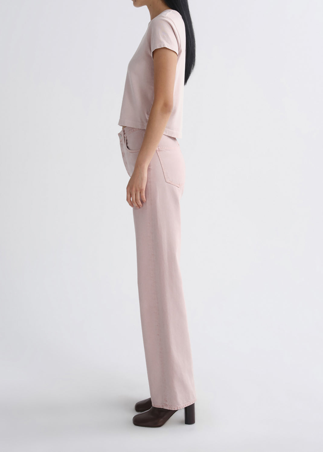 A model shown in profile wearing a light pink short-sleeve crop top and matching wide-leg trousers, paired with dark brown block-heel shoes, against a grey background.