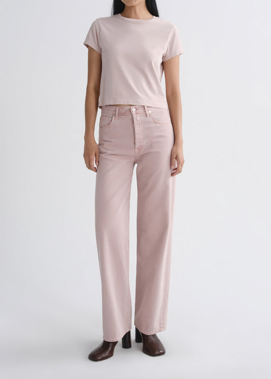 A model wearing a light pink short-sleeve crop top and matching wide-leg jeans, paired with dark brown heeled shoes, against a grey background.