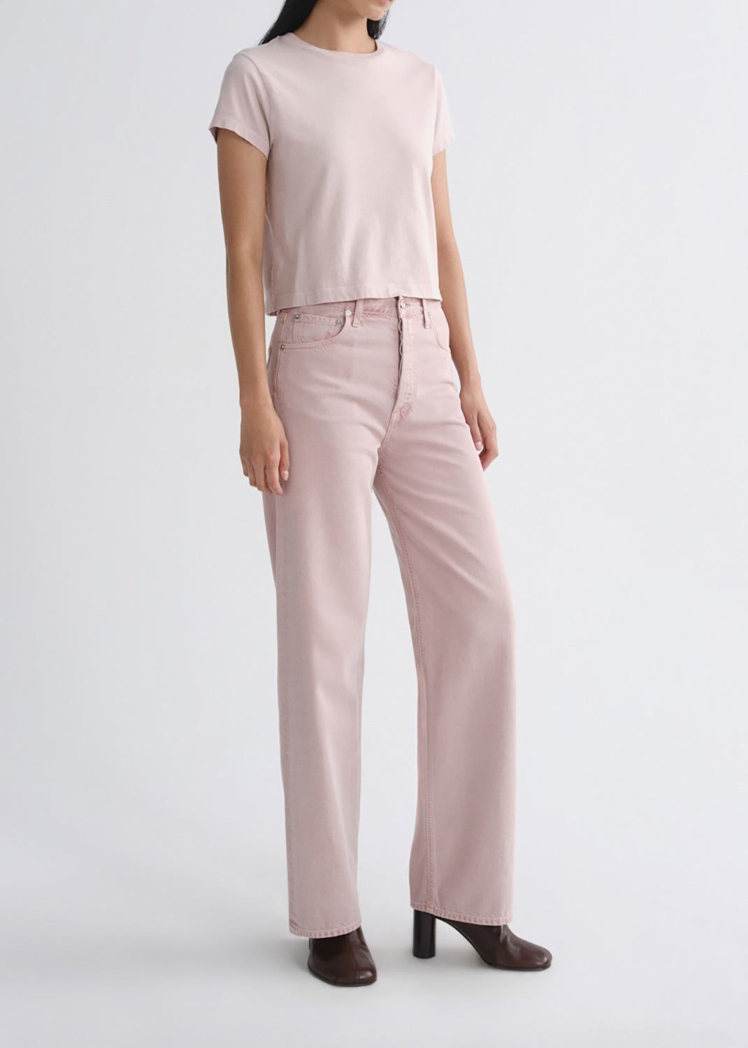 A model wearing a light pink short-sleeve crop top and matching light pink wide-leg jeans, paired with dark brown heeled shoes, against a grey background.