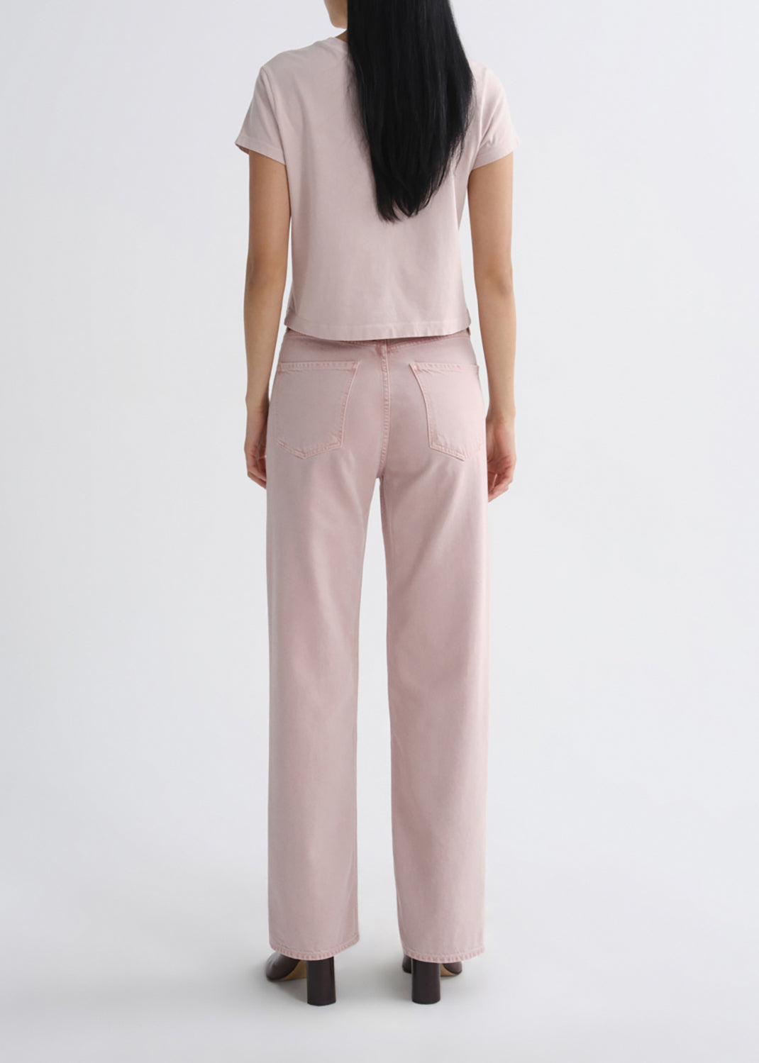 A model viewed from the back wearing a light pink short-sleeve crop top and matching light pink wide-leg jeans, paired with dark brown block-heel shoes, against a grey background.
