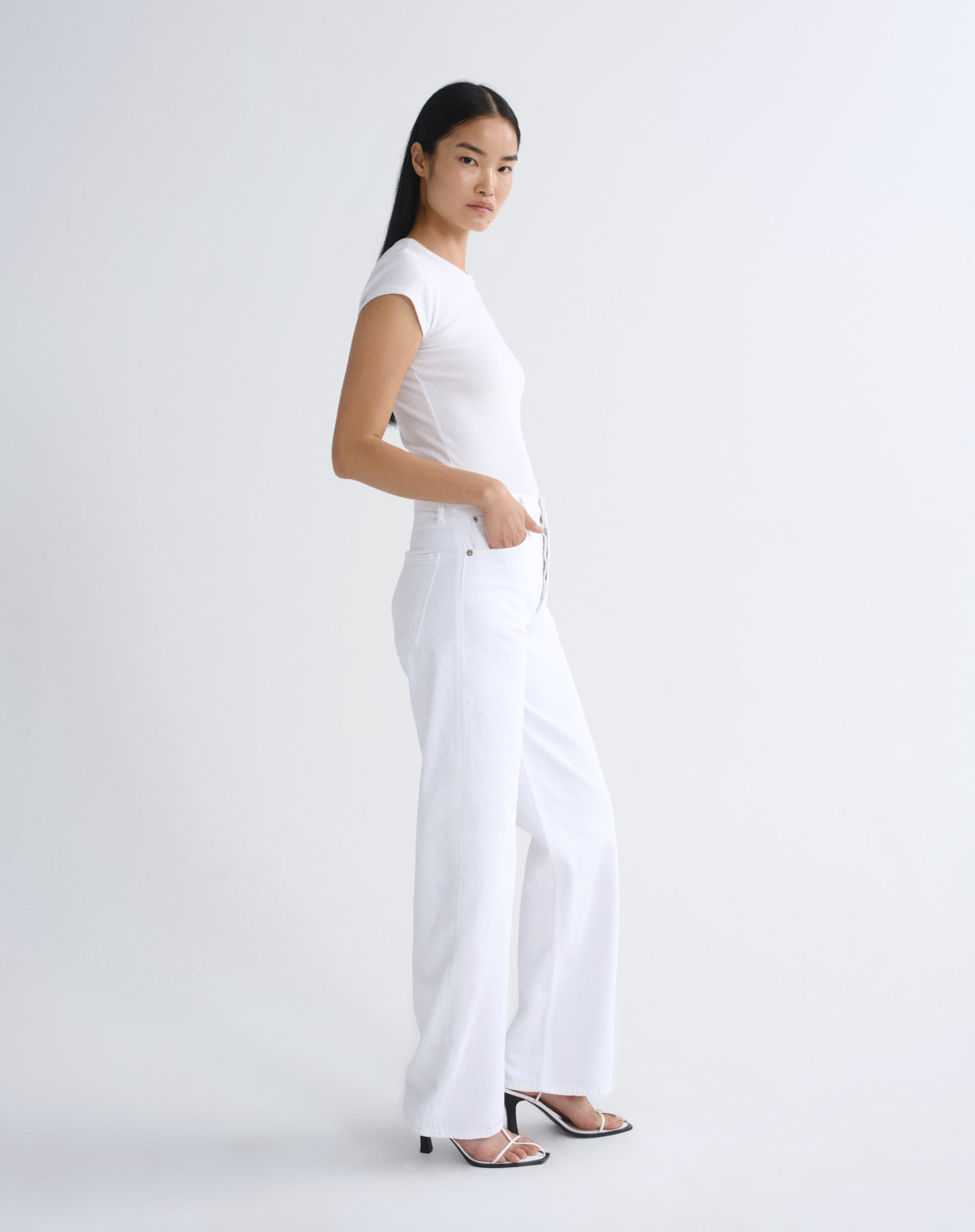 A woman modeling a white short-sleeve top and white wide-leg trousers, standing in profile against a clean white studio background.