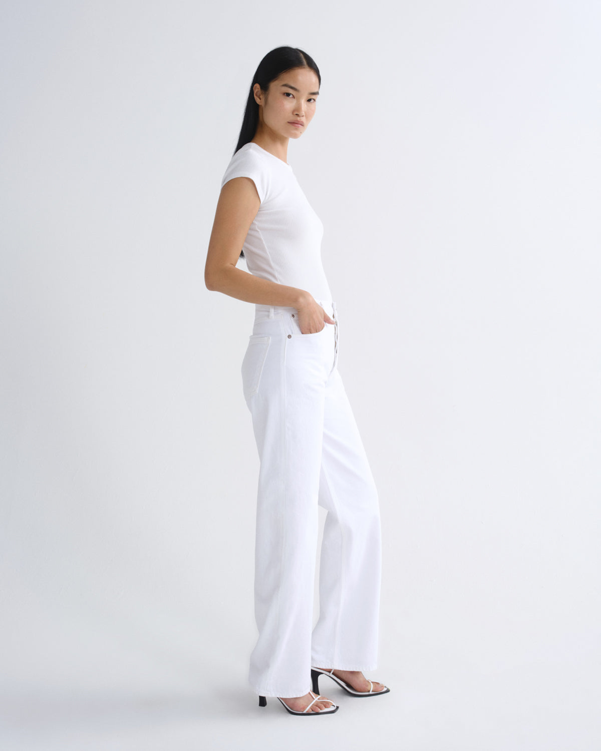 A woman modeling a white short-sleeve crewneck top and matching white wide-leg trousers, paired with black strappy high-heeled sandals, against a seamless white studio background.