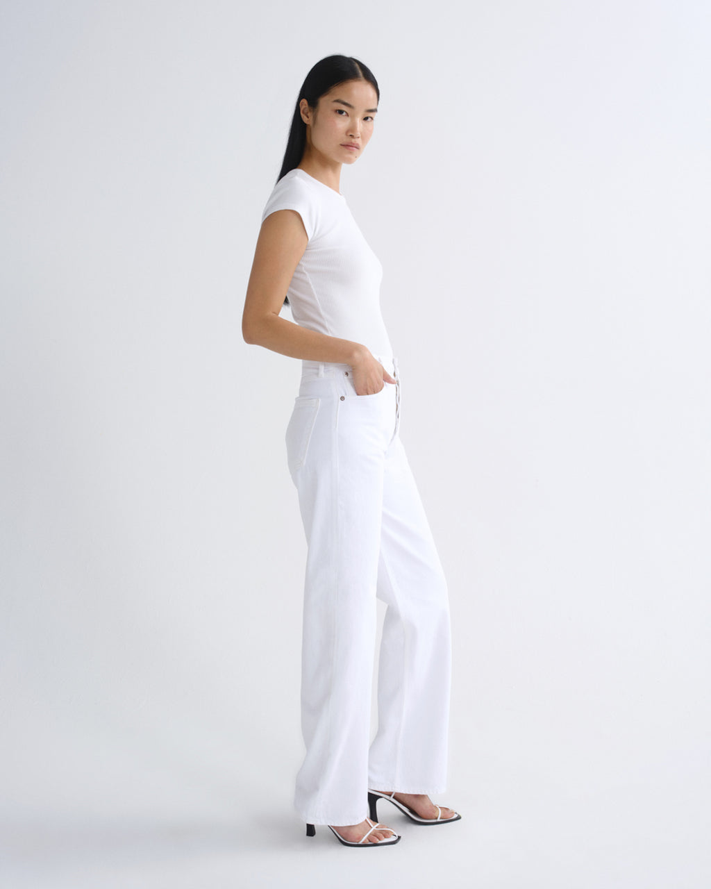 A woman modeling a white short-sleeve crewneck top and matching white wide-leg trousers, paired with black strappy high-heeled sandals, against a seamless white studio background.
