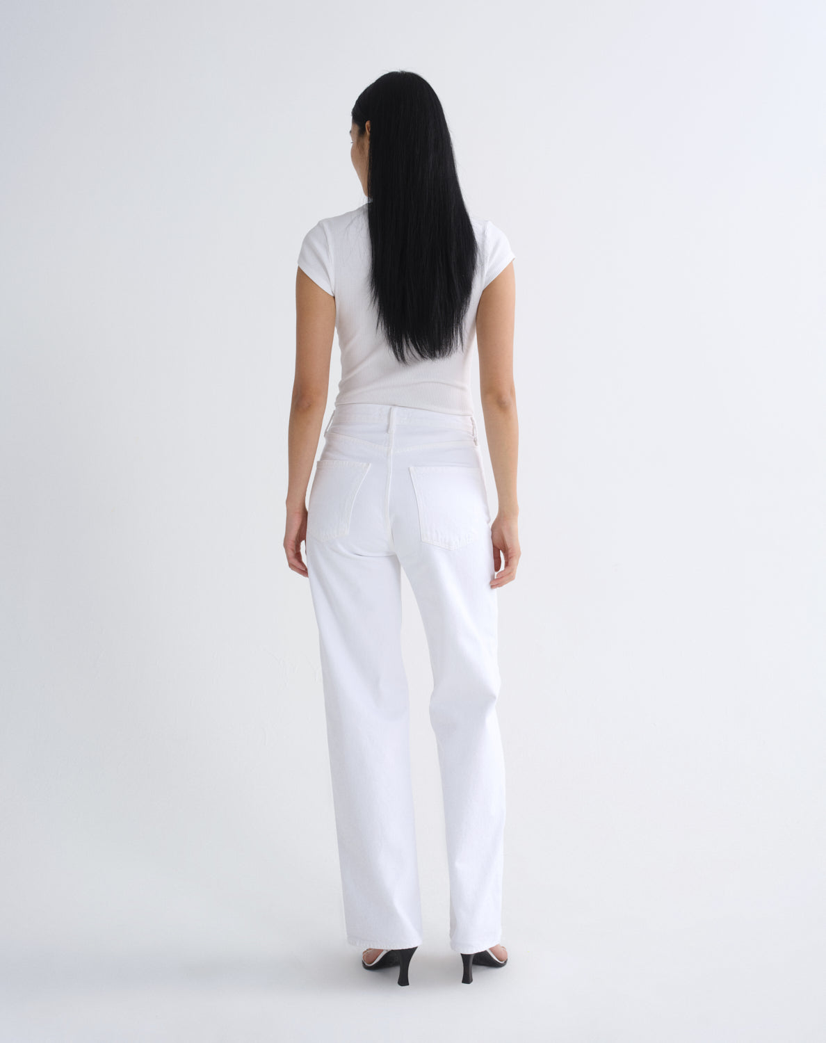 A woman with long black hair is shown from behind wearing a white short-sleeve top and white wide-leg jeans, standing against a plain light gray studio background.