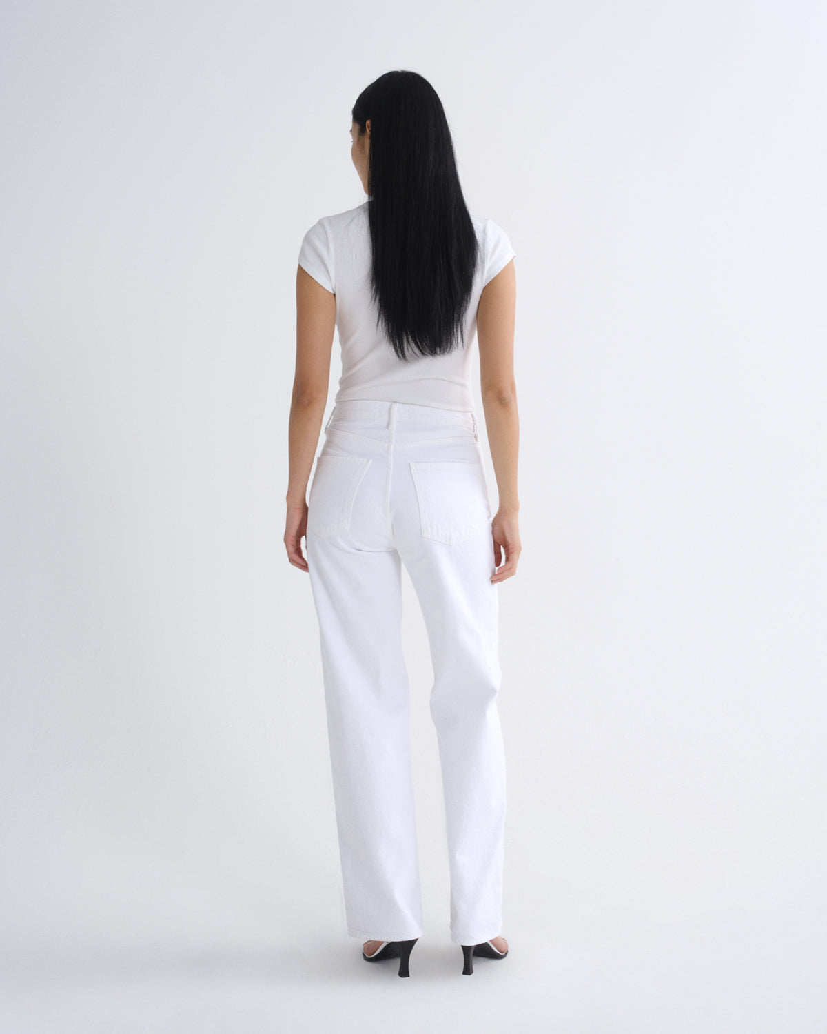 A woman with long black hair is shown from behind wearing a white short-sleeve top and white wide-leg jeans, standing against a plain light gray studio background.