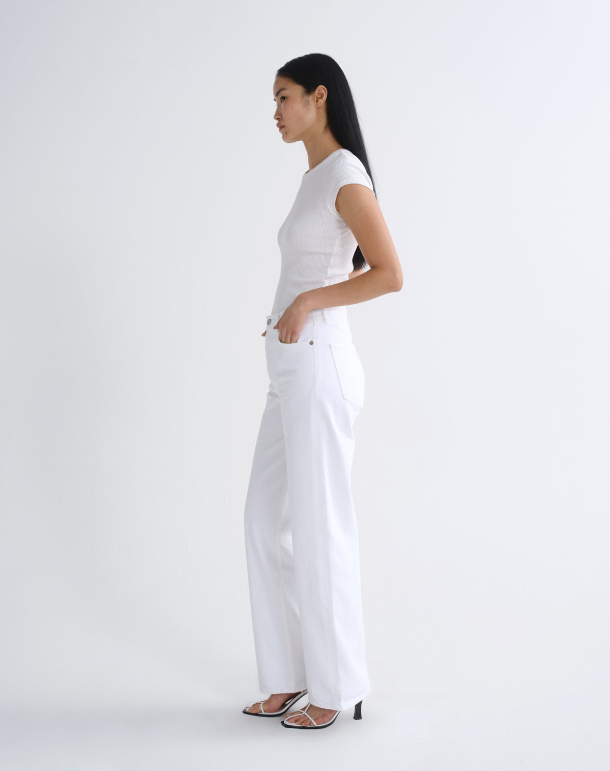 A woman in a white short-sleeve top and white wide-leg pants stands in profile against a clean white studio background, wearing black heeled sandals.