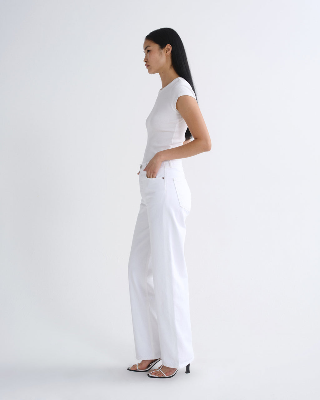 A woman in a white short-sleeve top and white wide-leg pants stands in profile against a clean white studio background, wearing black heeled sandals.