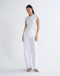 A woman modeling a white short-sleeve crewneck top and white wide-leg jeans with black strappy heels, standing against a seamless white studio background.