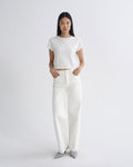 A model stands against a light gray studio background wearing a white short-sleeve cropped T-shirt, white wide-leg pants, and gray pointed shoes.