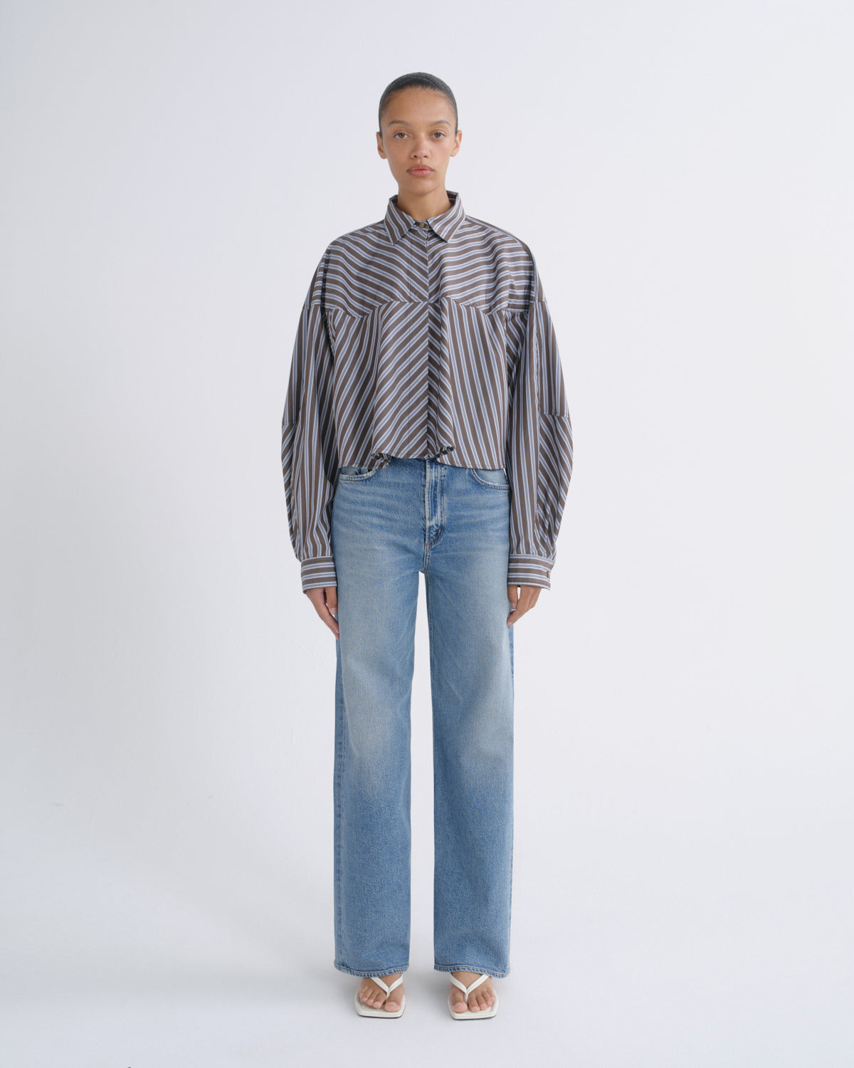A model stands against a light gray studio background wearing a gray and black striped button-up shirt with wide sleeves, paired with light-wash blue wide-leg jeans and white sandals.