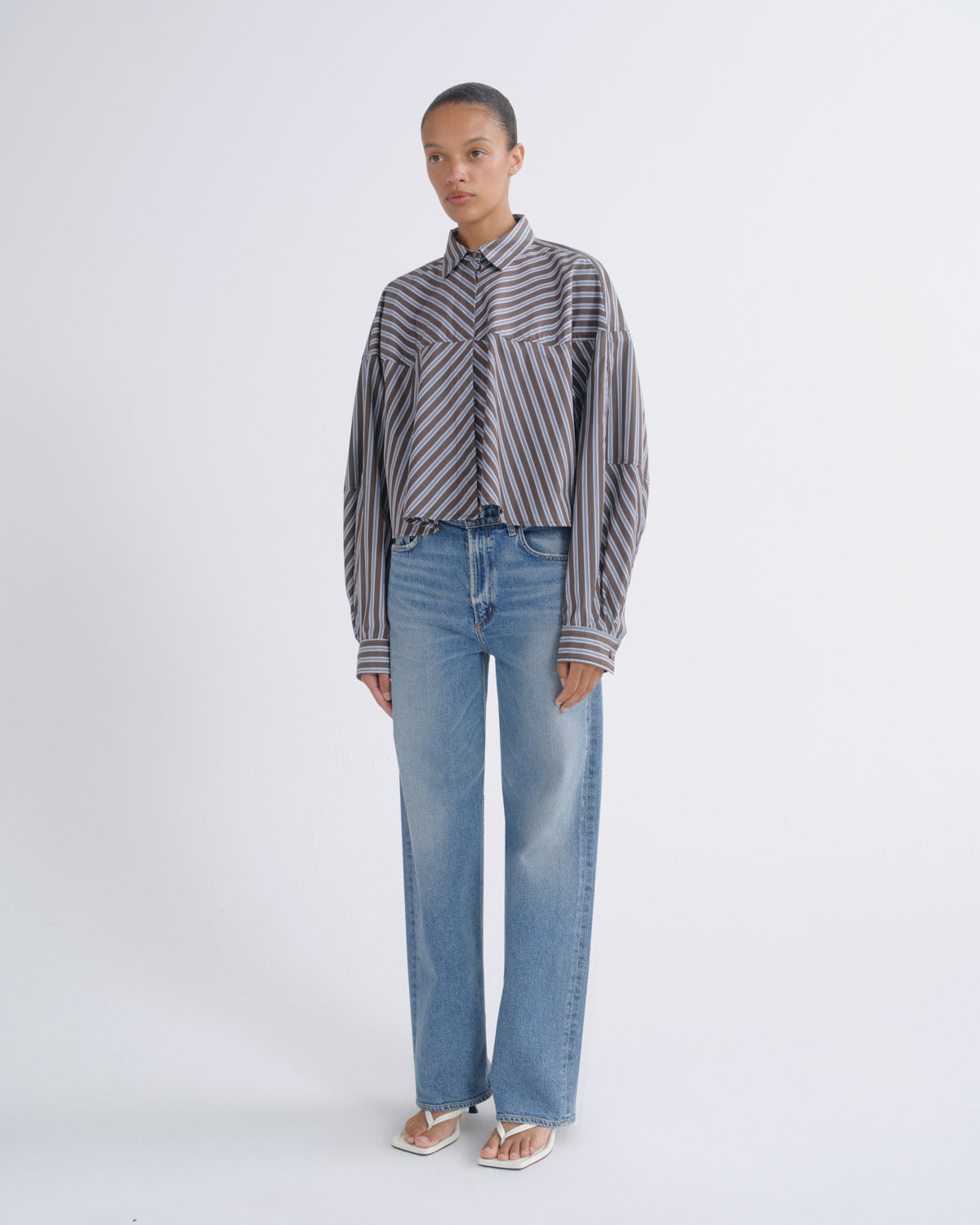 A model stands against a light gray studio background wearing a cropped, long-sleeve button-up shirt with vertical gray and white stripes, paired with light-wash blue jeans and white sandals.