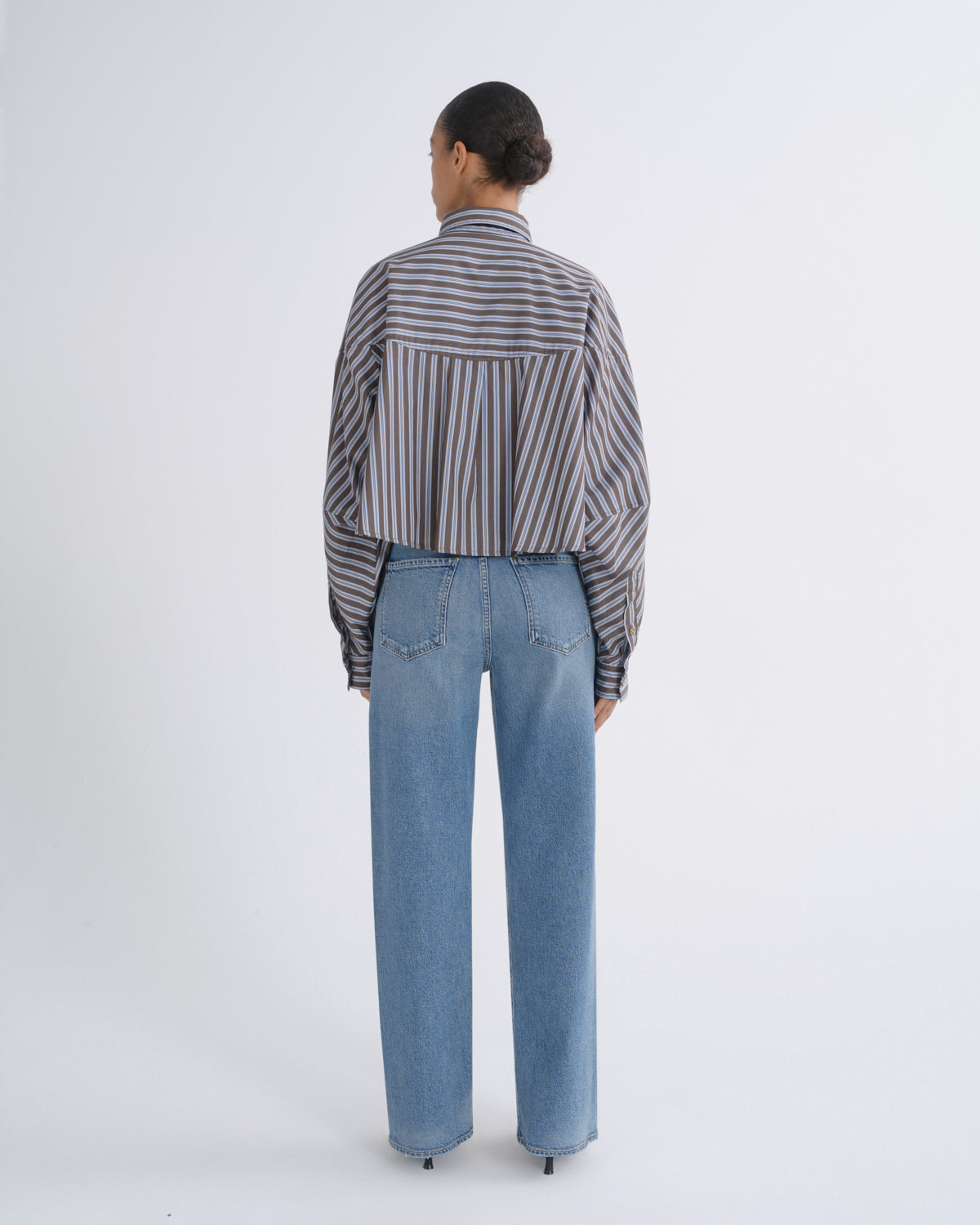 A model viewed from the back wearing a cropped, long-sleeve striped button-up shirt in gray, white, and dark brown, paired with light-wash blue jeans, against a plain light gray studio background.