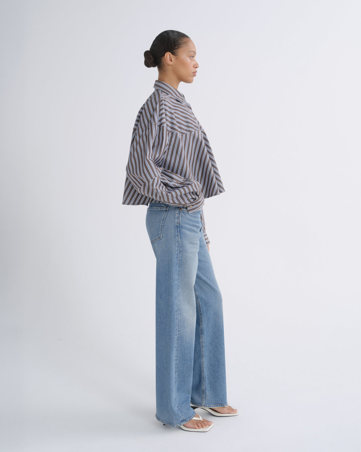 A woman stands in profile wearing a cropped, oversized button-up shirt with vertical gray and white stripes, light-wash wide-leg jeans, and flat sandals against a plain light gray studio background.
