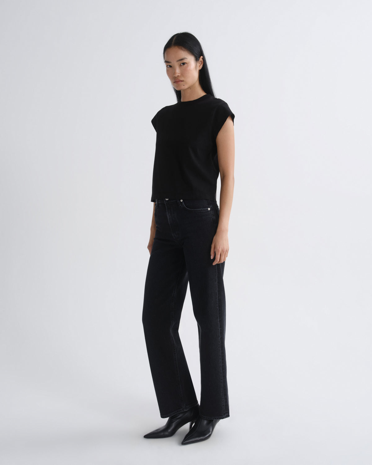 A model stands in a studio wearing a black sleeveless top, black wide-leg jeans, and black pointed-toe heels against a light gray background.